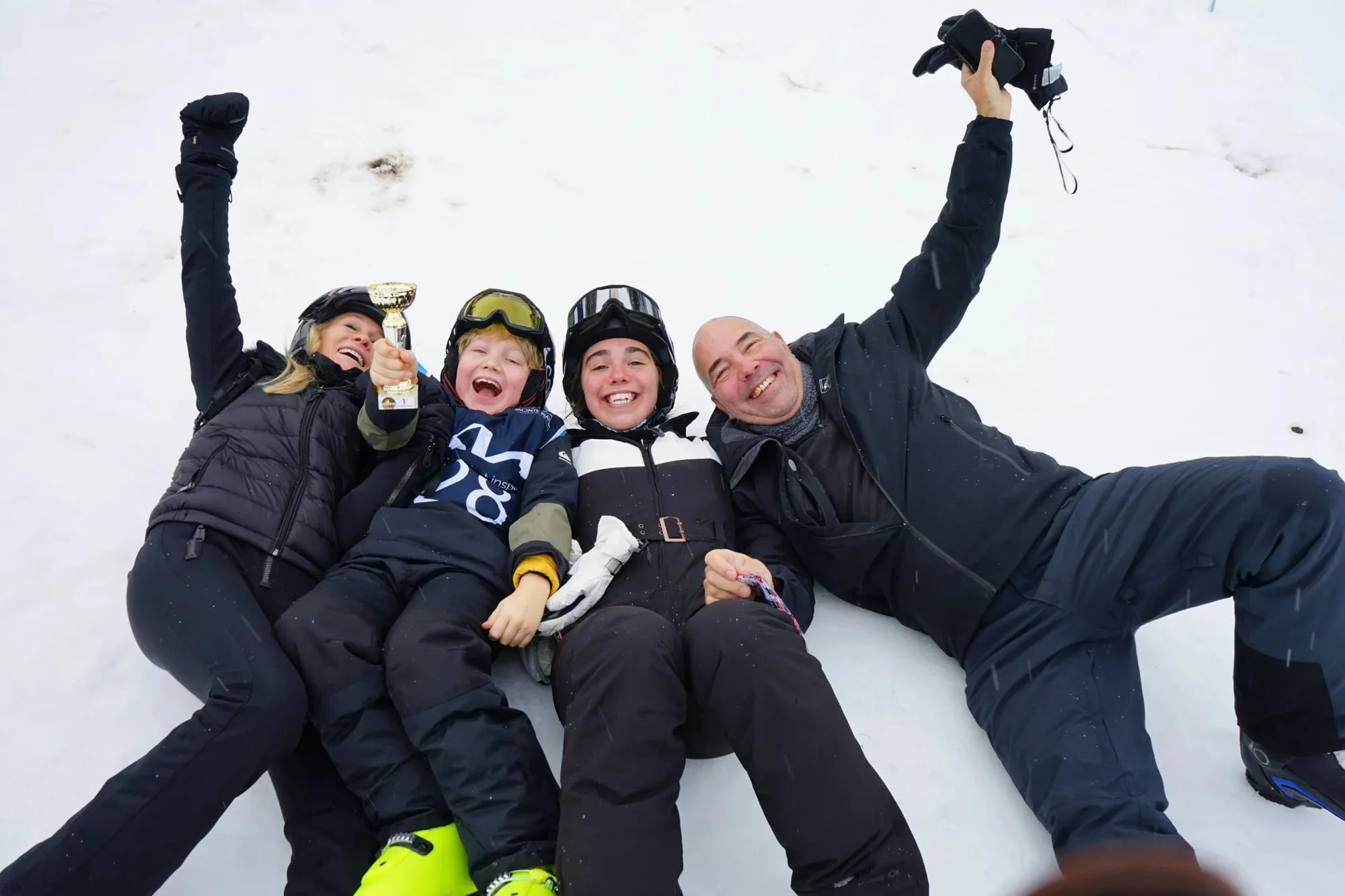Group of four people lying on snow, celebrating. One child holding a trophy, all wearing winter gear and helmets, smiling and enjoying snowy outdoors. Very happy family ski holiday in courmayeur