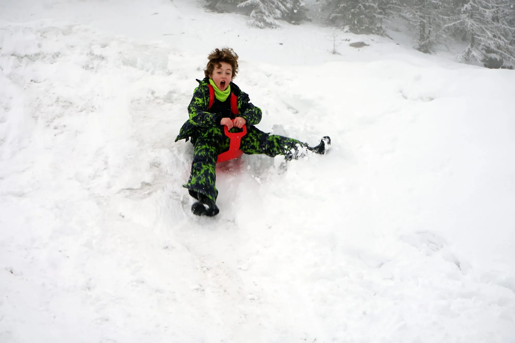 A young child in winter gear sitting on a red sled in the snow with a surprised expression on his face, surrounded by snow-covered trees and landscape.